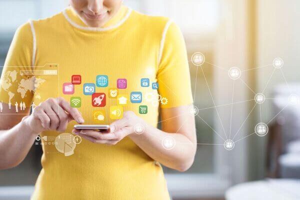 Joven sonriendo sostiene en sus manos un móvil desde donde salen imágenes de las diferentes redes sociales. Claramente aplicando una estrategia de marketing